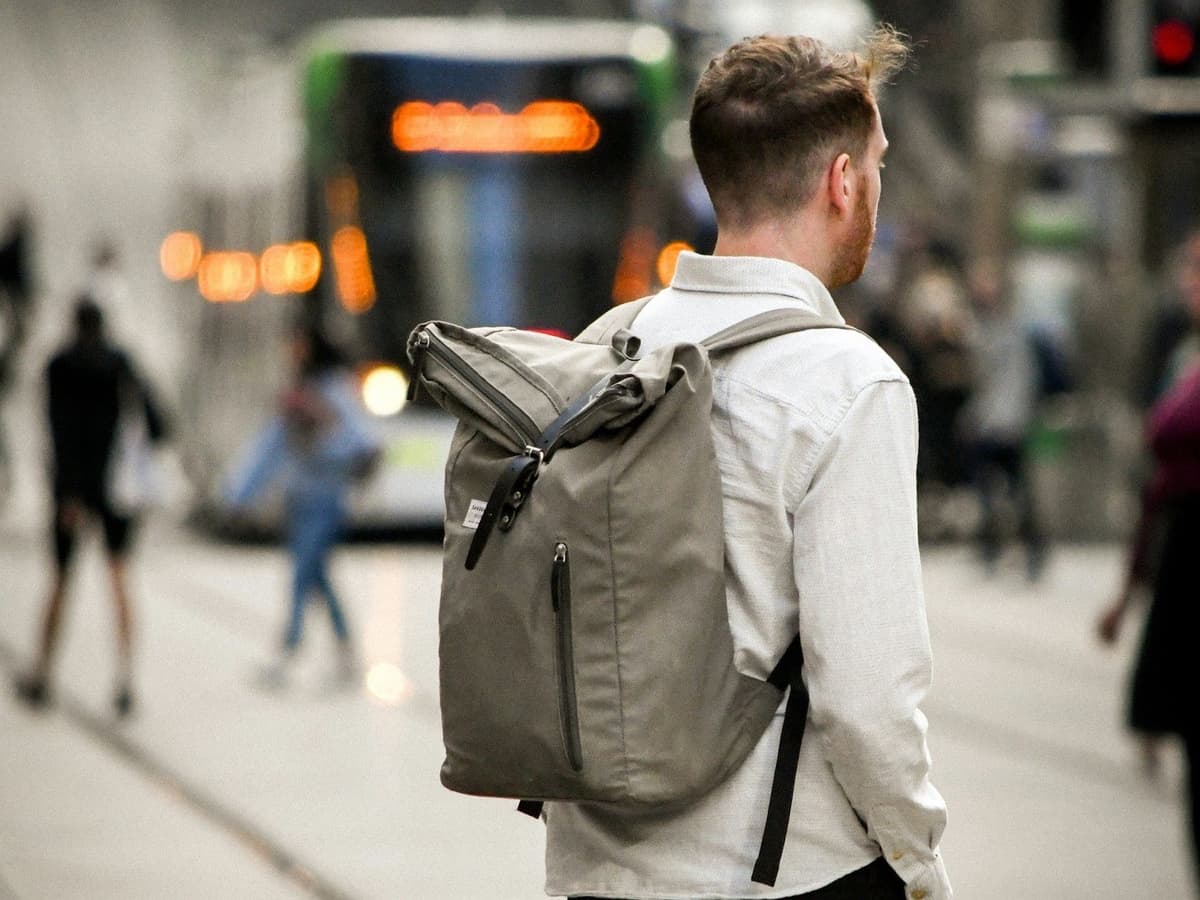 A commuter seen from behind wearing a grey laptop backpack, waiting to cross as a tram passes