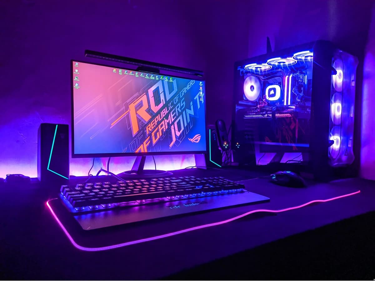 A gaming monitor showing an ROG wallpaper beside an RGB-lit desktop PC on a dark desk