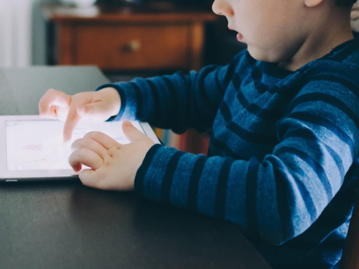 A young boy in a striped jumper tapping the screen of a white tablet at a table