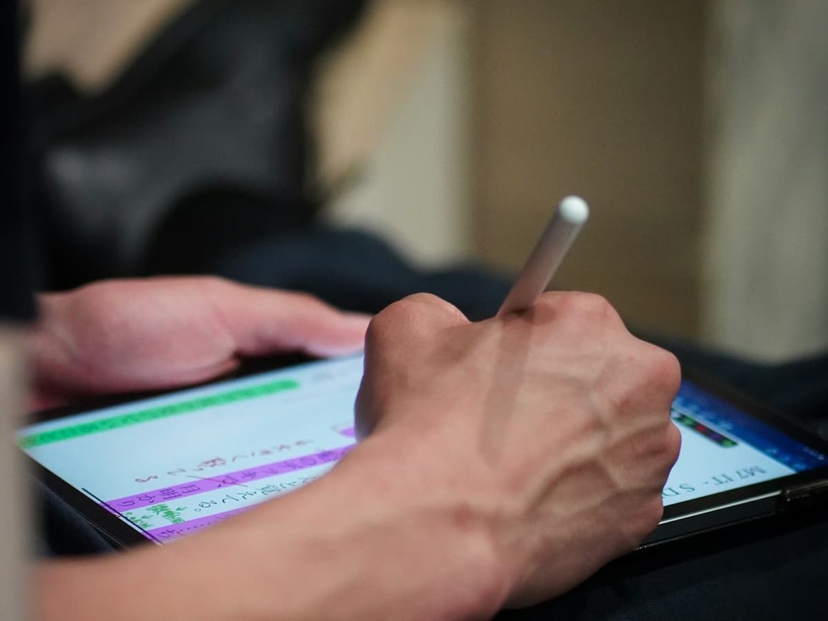 A hand holding a stylus writing highlighted handwritten notes on a tablet held in the lap