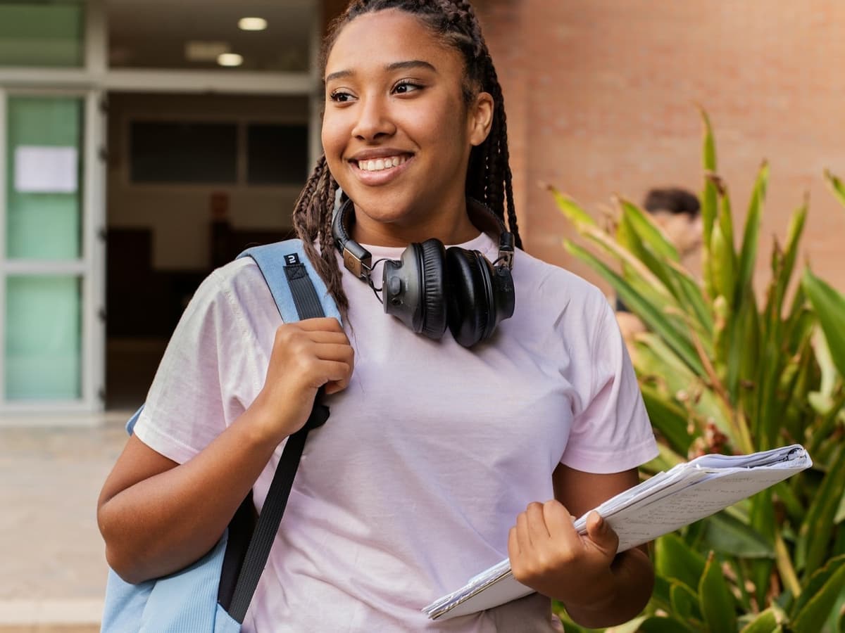 A smiling student on campus with a blue backpack over one shoulder, holding a stack of lecture notes