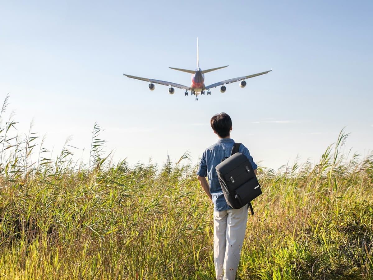 A traveller seen from behind in a field wearing a structured black travel backpack as a jet comes in to land