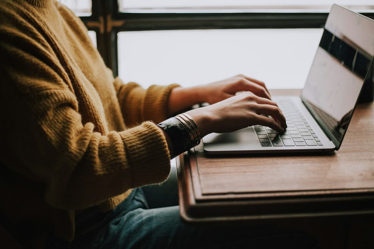A student typing on a laptop at a wooden desk beside a window