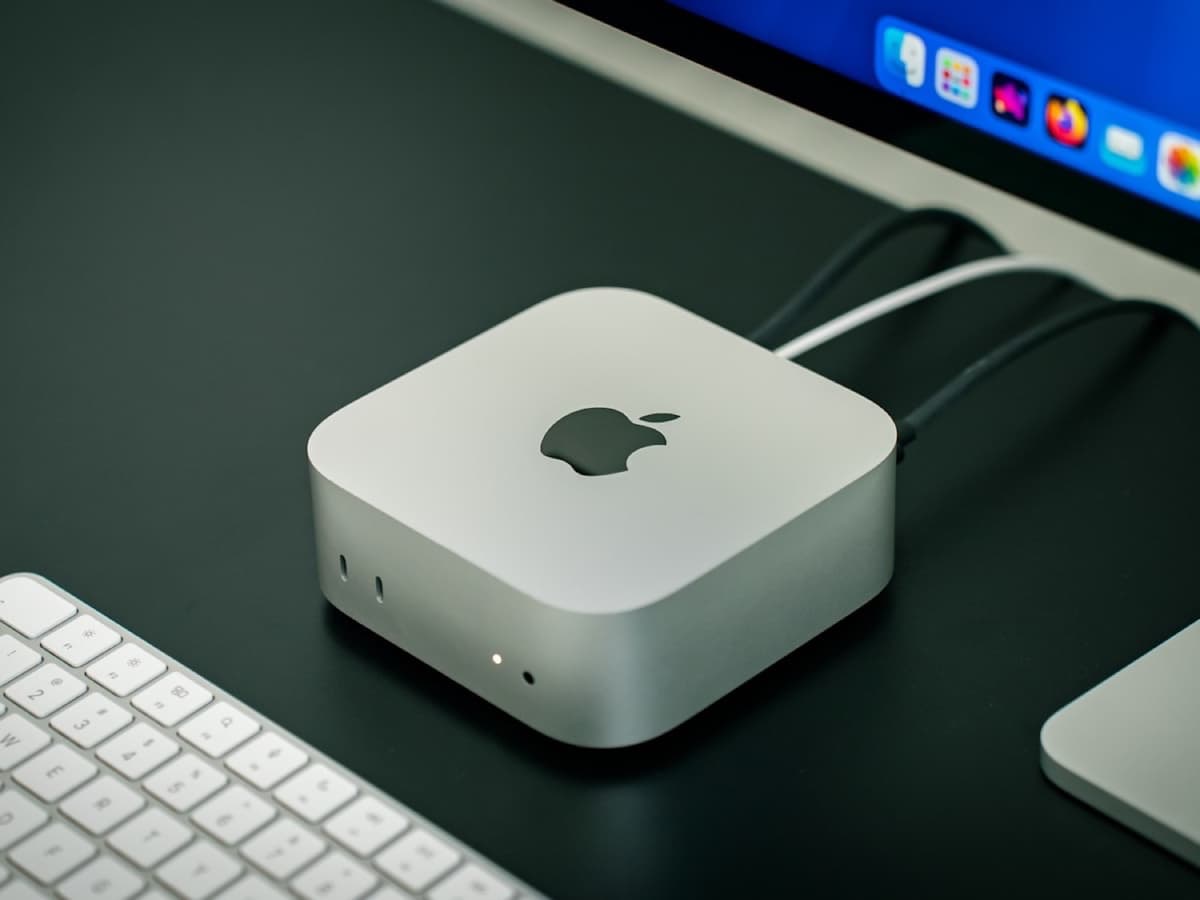 A small silver mini PC on a desk beside a wireless keyboard, a trackpad and a monitor
