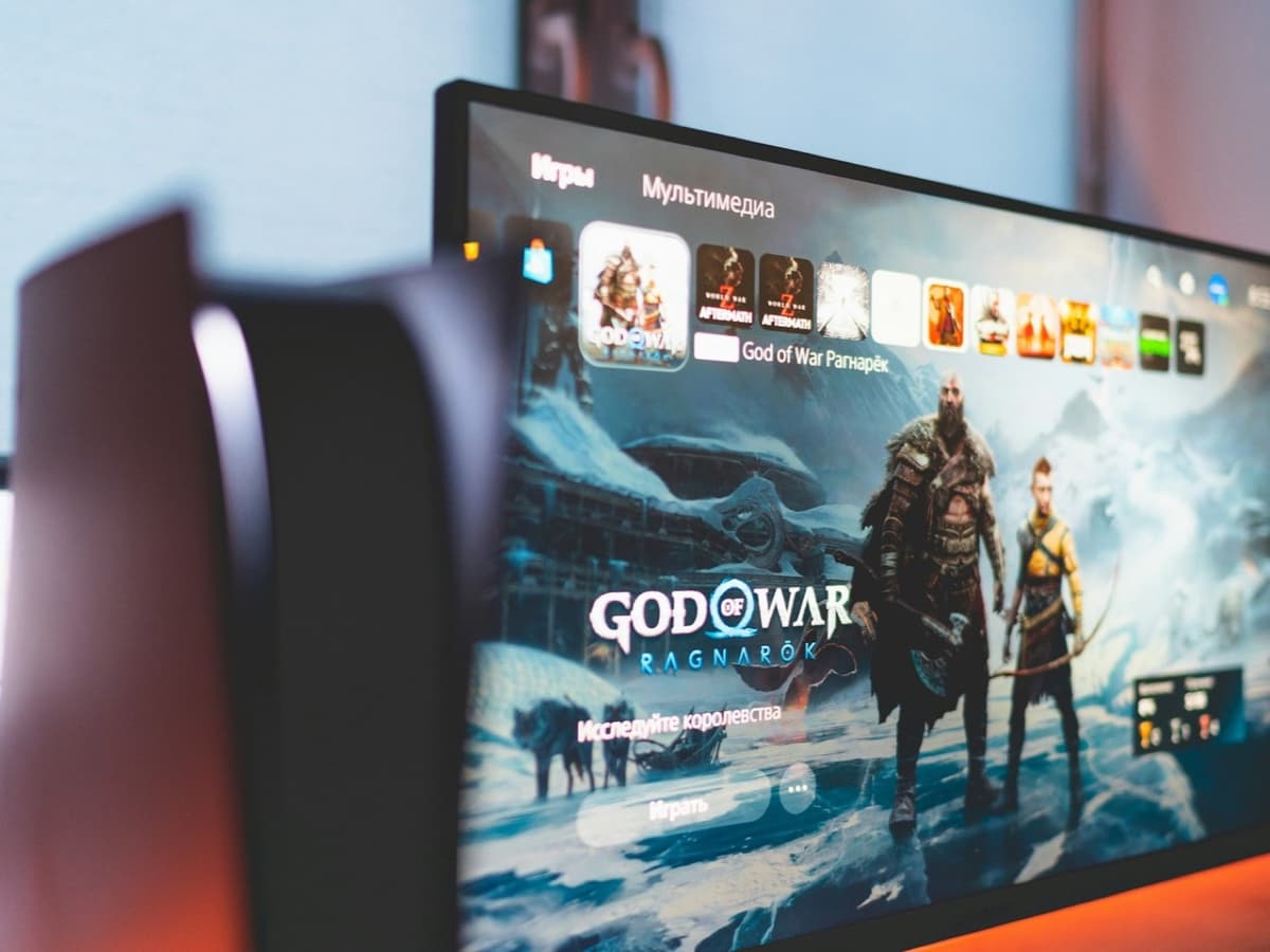 A white PlayStation 5 console beside a monitor displaying the PS5 home screen
