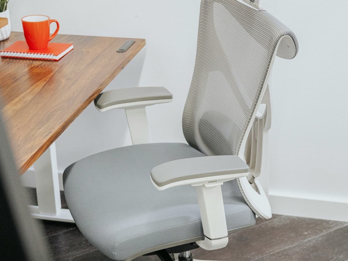 A grey mesh-backed ergonomic office chair with adjustable arms pulled up to a wooden desk