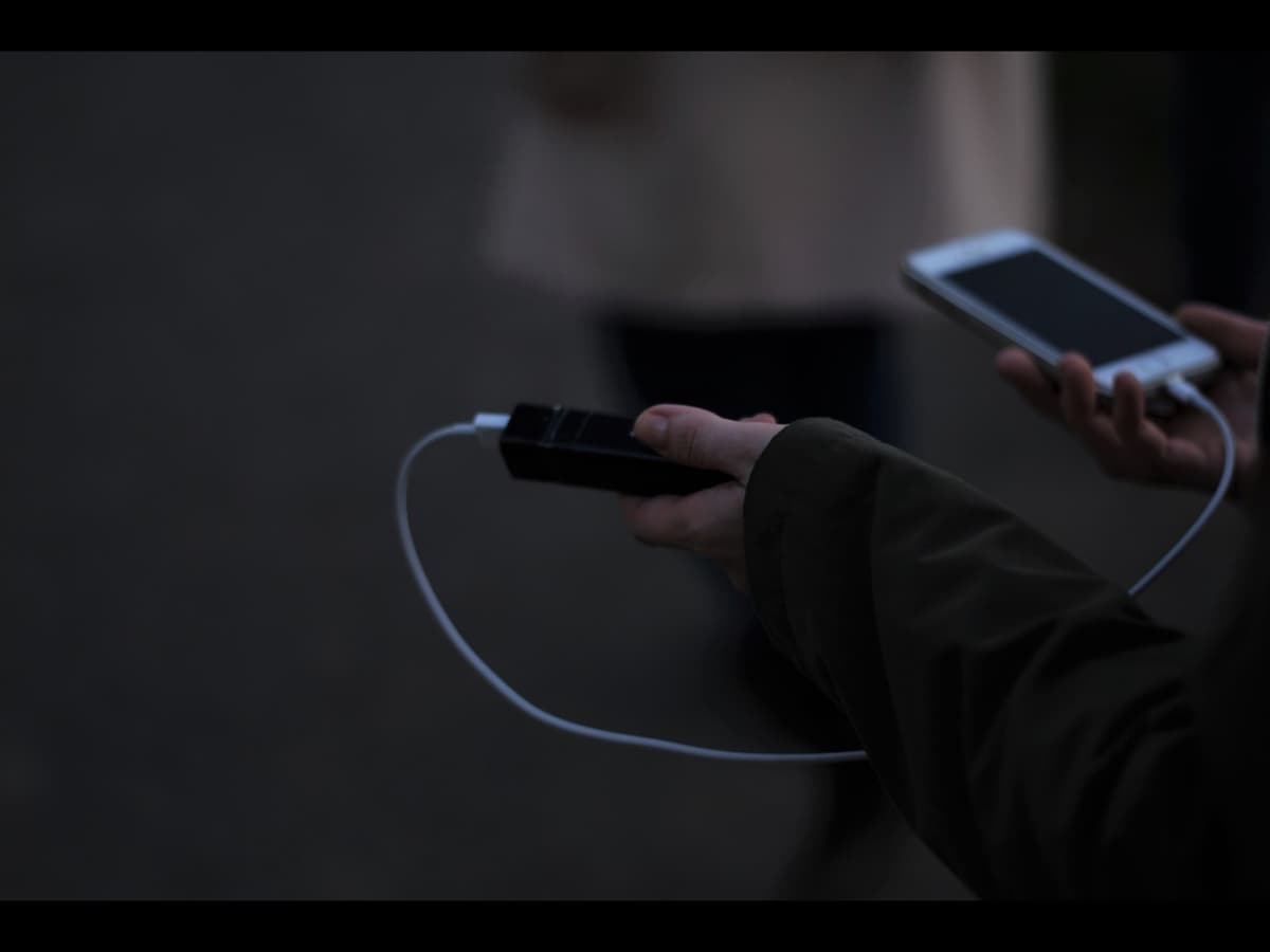 A person outdoors holding a small black power bank charging a phone by cable