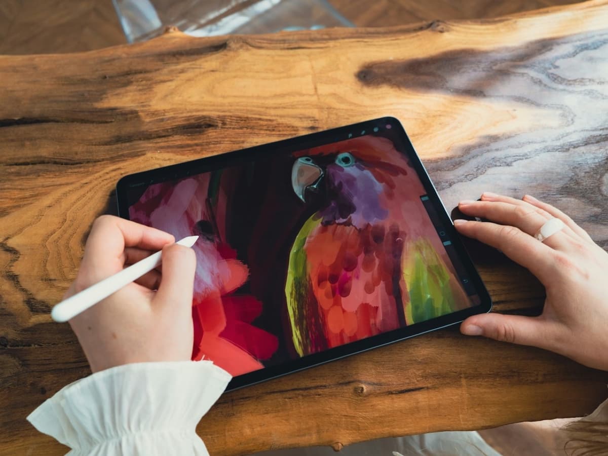 A hand using a white stylus to paint a colourful parrot on a tablet resting on a wooden table